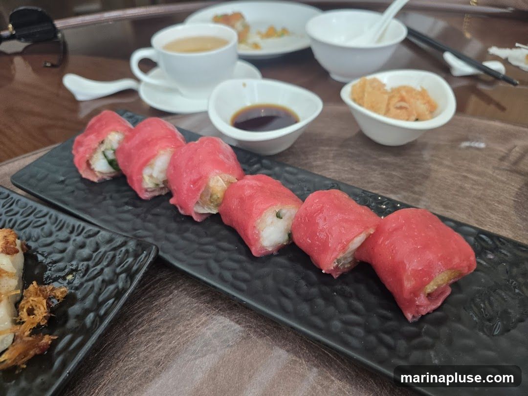 A close-up shot of a dim sum platter featuring several pieces of sushi-like rolls with pink wrappers and white fillings, served on a black textured plate. In the background, there are small white bowls containing soy sauce and crispy fried items, a cup of tea, and chopsticks. Another black plate with a partially eaten dish is visible on the left.