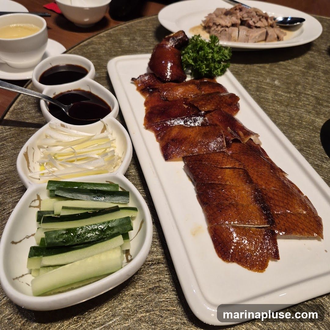A platter of sliced Peking duck with crispy skin is served with accompaniments including sliced cucumber, scallions, and dipping sauces. A bowl of soup and another dish of sliced meat are also visible on the table.