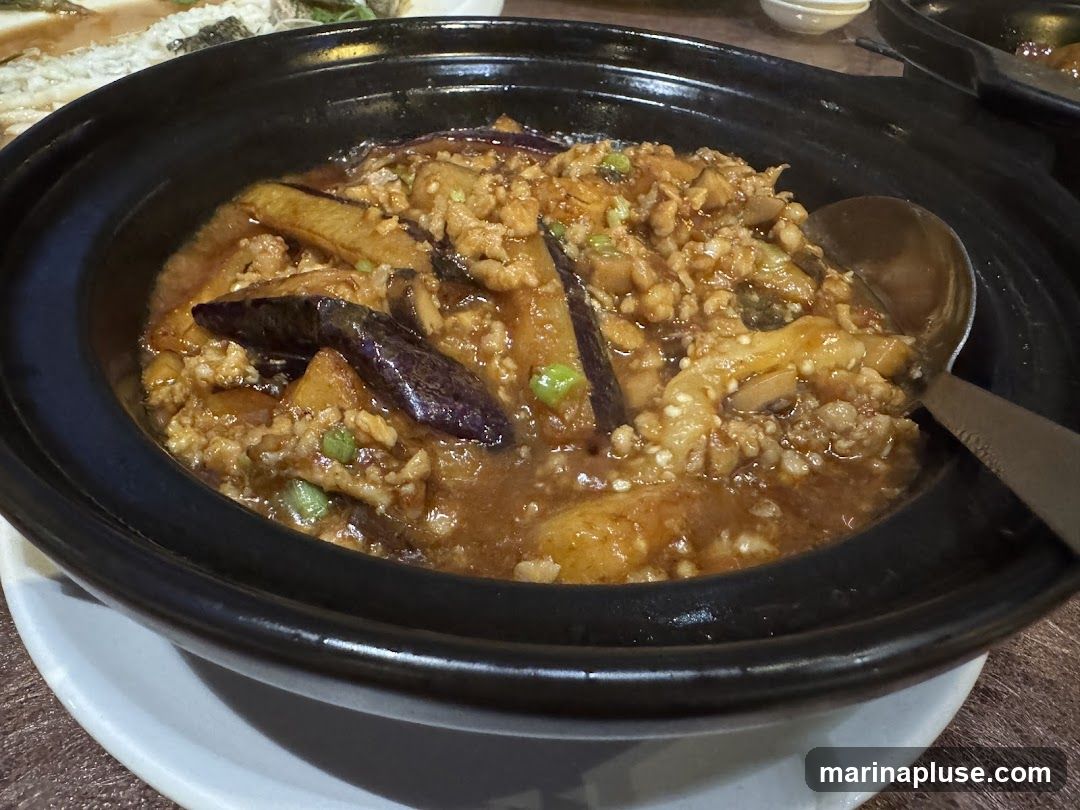 A close-up shot of a dish featuring braised eggplant and minced meat in a savory sauce, served in a black ceramic pot. A large spoon is partially submerged in the dish, ready to serve.