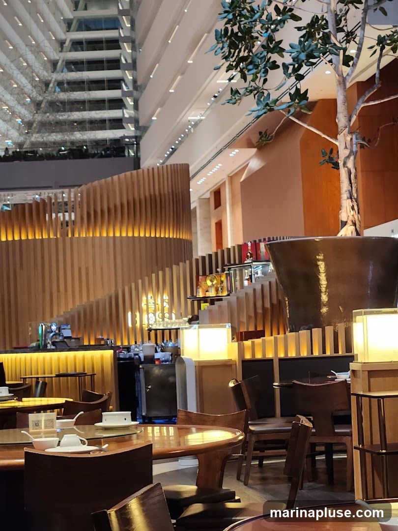 The image shows the interior of a restaurant with a prominent bar area and dining tables. A large, decorative tree in a pot is visible on the right. The architecture features a multi-level atrium with wooden slatted accents and warm lighting. Tables are set with white teacups and saucers. The bar area has bottles and glasses displayed.