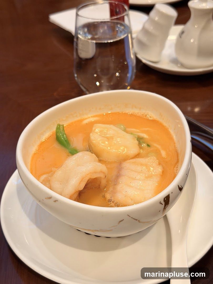 A close-up shot of a bowl of seafood soup with large pieces of shrimp and fish in a creamy orange broth. A glass of water and salt and pepper shakers are visible in the background.