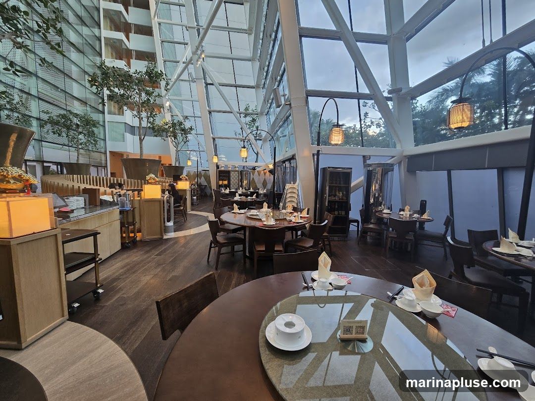 A spacious restaurant interior with a modern, geometric glass ceiling and wooden floors. Several round dining tables are set with white tableware and napkins. Potted trees are placed throughout the dining area. In the background, a buffet station is visible with various food items and serving dishes. The overall ambiance is elegant and contemporary.