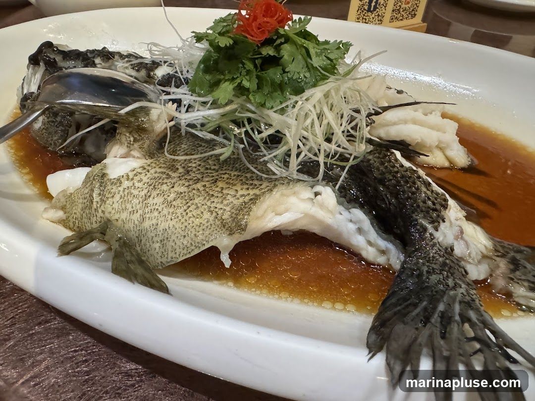 A whole steamed fish served on a white platter with a savory sauce. The fish is garnished with shredded scallions and cilantro, topped with a red garnish. A spoon is placed in the fish's head.