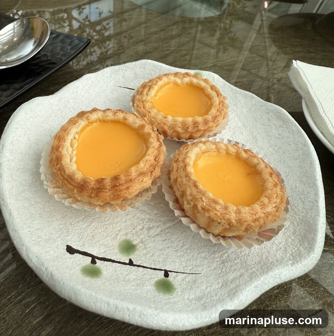 Three golden-brown egg tarts with a smooth, bright yellow custard filling are arranged on a white, textured plate with a minimalist branch and leaf design. A spoon and a black rectangular dish are visible in the upper left corner, and a white napkin is on the right. The table surface has a reflective, marbled pattern.