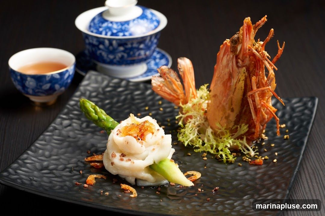 A close-up shot of a gourmet seafood dish served on a textured black plate. The dish features a large, crispy fried prawn and a delicate swirl of white seafood mousse wrapped around an asparagus spear, garnished with small dried shrimp and herbs. To the left, a traditional blue and white porcelain tea cup filled with amber liquid and a matching lidded teapot are visible, suggesting a tea pairing. The dark background enhances the presentation of the food.