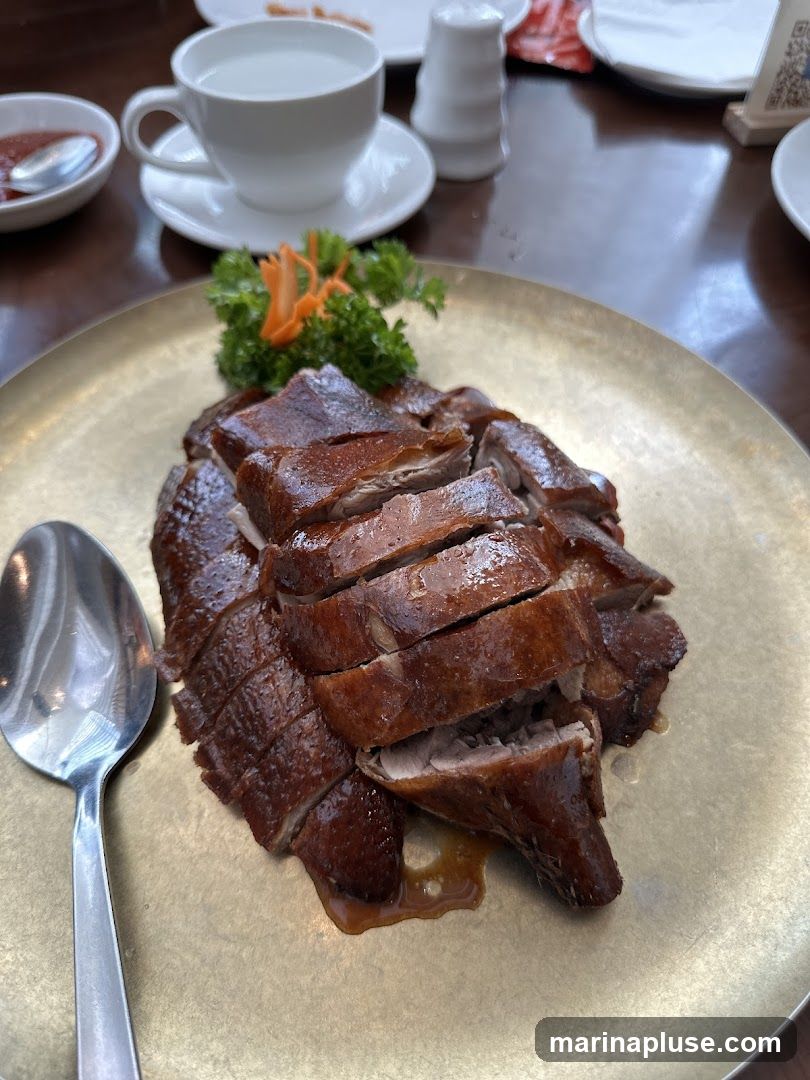 A close-up shot of a sliced roasted duck dish served on a golden plate. The duck has crispy, glistening skin and is garnished with parsley and shredded carrots. A spoon rests on the left side of the plate. In the background, a cup of water and a small dish of sauce are visible on a wooden table.