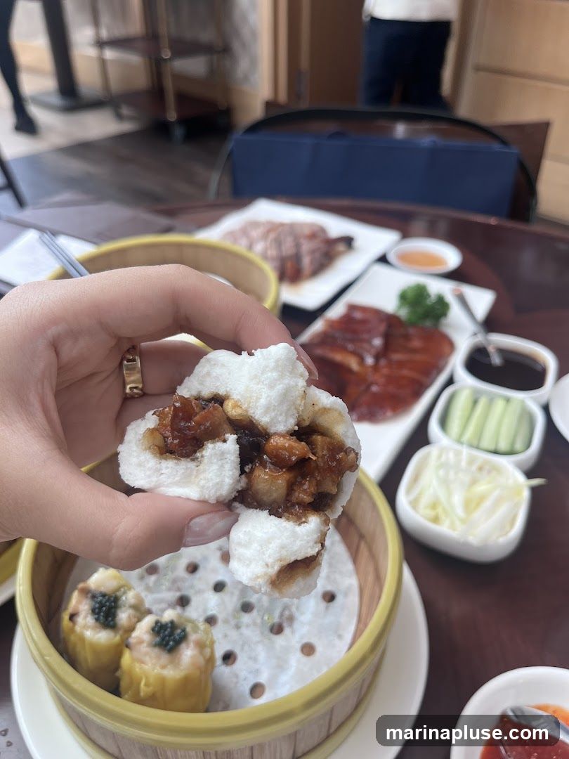A hand holds a steamed bun filled with meat. In the background, a table is set with various dishes, including roasted duck, siu mai dumplings, sliced cucumbers, and dipping sauces. The setting appears to be a restaurant.