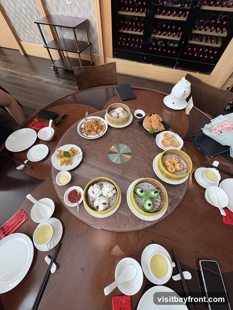 A round table laden with various dim sum dishes, including steamed dumplings, fried items, and small pastries. Several cups of tea and dipping sauces are also visible. In the background, a wine rack filled with bottles and a serving cart can be seen.