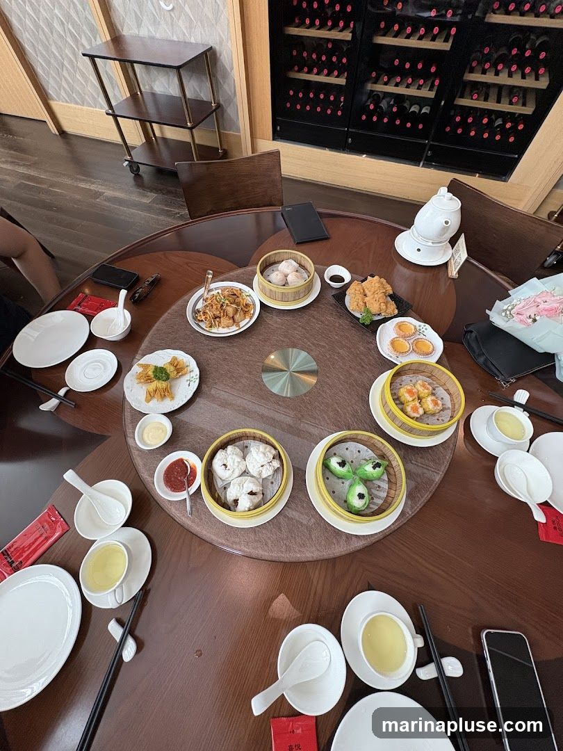 A round table laden with various dim sum dishes, including steamed dumplings, fried items, and small pastries. Several cups of tea and dipping sauces are also visible. In the background, a wine rack filled with bottles and a serving cart can be seen.