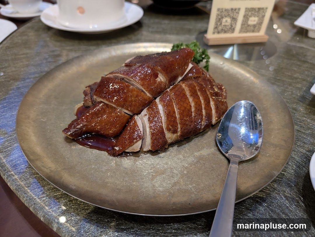 A plate of sliced roasted duck with a glossy glaze, served on a metallic plate. A spoon rests to the right of the plate. In the background, there are white teacups and saucers, and a wooden stand with QR codes.