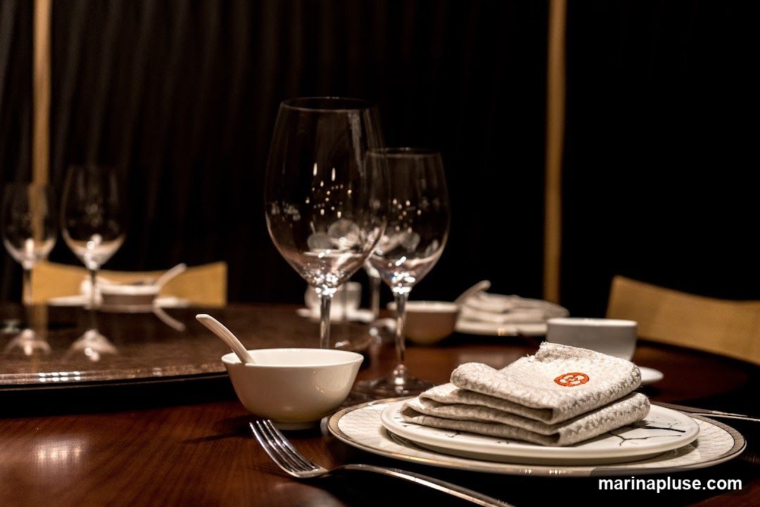 A formal table setting with wine glasses, plates, cutlery, and folded napkins. The napkins have an embroidered logo. The lighting is dim, creating an intimate atmosphere.