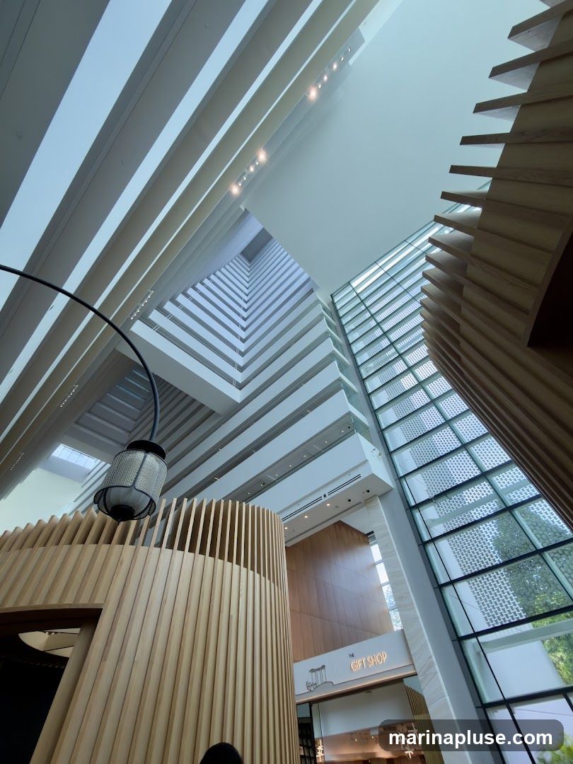 A low-angle shot looking up at the interior of a large, modern building. The architecture features multiple levels of balconies or walkways, a large glass facade, and wooden decorative elements. A sign for 'THE GIFT SHOP' is visible on one of the lower levels.