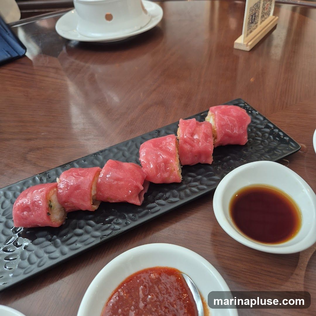 A close-up shot of a dim sum dish featuring several pieces of pink-colored dumplings, likely rice noodle rolls, served on a black textured plate. The dumplings are filled with a light-colored ingredient and appear to be garnished with something green. Two small white bowls are placed in front of the plate, one containing a dark soy sauce and the other a red chili sauce. A white teacup and saucer are visible in the blurred background, along with a small wooden stand displaying a menu or advertisement. The setting appears to be a restaurant table with a polished wooden surface.