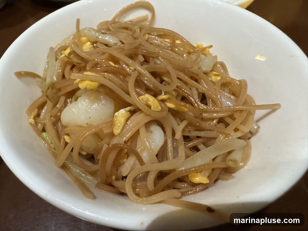 A close-up shot of a white bowl filled with thin noodles, scrambled egg, and pieces of scallop. The noodles are coated in a light brown sauce.