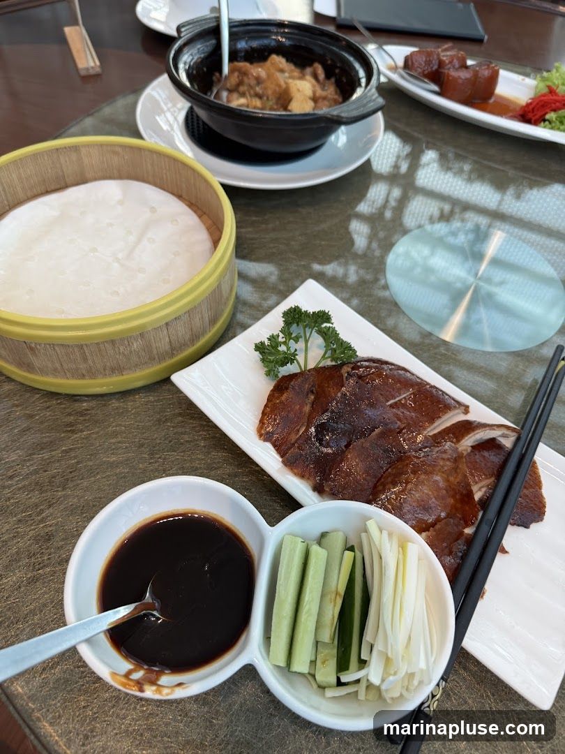 A table setting with a Peking duck meal. The meal includes sliced Peking duck, a bamboo steamer filled with thin pancakes, a small bowl of dark sauce, and a dish with sliced cucumber and scallions. In the background, there is a pot of stew and another dish with meat.