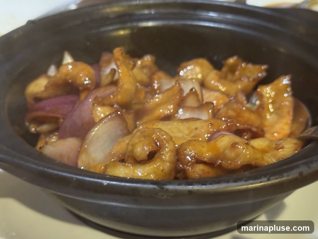A close-up shot of a stir-fried dish served in a dark, round bowl. The dish appears to contain pieces of meat or tofu coated in a glossy, brown sauce, mixed with chunks of red onion. The food is steaming, suggesting it is freshly served.
