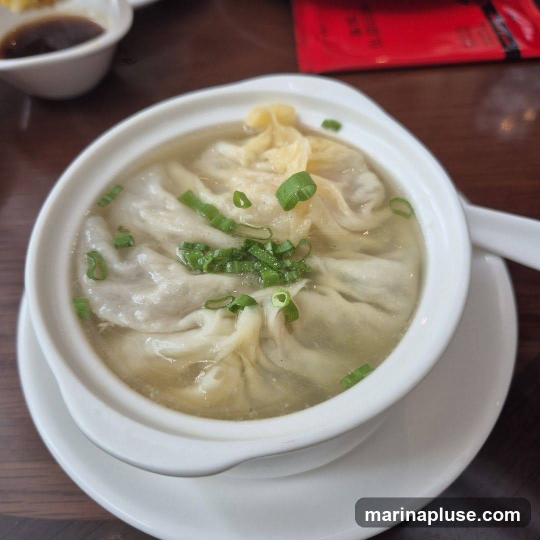 A close-up shot of soup dumplings in a white bowl, garnished with chopped green onions. A small dish of dipping sauce is visible in the upper left corner.