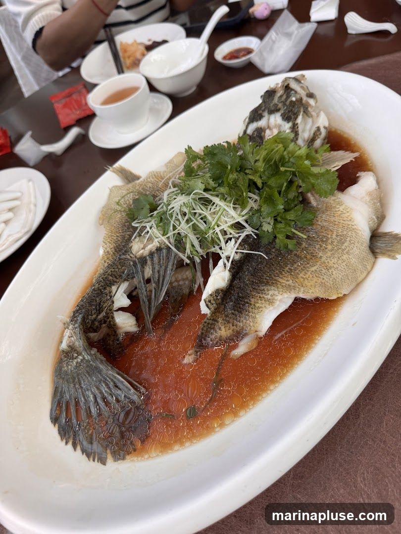 A whole steamed fish served on a white oval platter, garnished with fresh cilantro and thinly sliced scallions. The fish is covered in a savory brown sauce with visible oil droplets. In the background, there are white bowls, a cup of tea, and chopsticks, suggesting a meal setting.