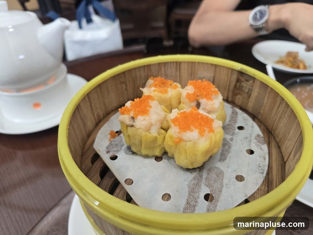 A close-up shot of dim sum dumplings in a bamboo steamer basket. The dumplings are topped with bright orange roe. In the background, a white teapot and a person wearing a watch are visible.