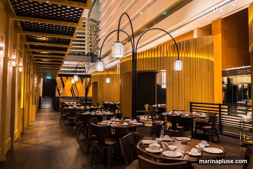 The interior of a restaurant with a warm, modern Asian-inspired design. Wooden accents, ambient lighting, and elegant table settings create a sophisticated dining atmosphere. Tables are set with white tableware, and decorative lanterns hang from arched fixtures. In the background, a "GIFT SHOP" sign is visible.