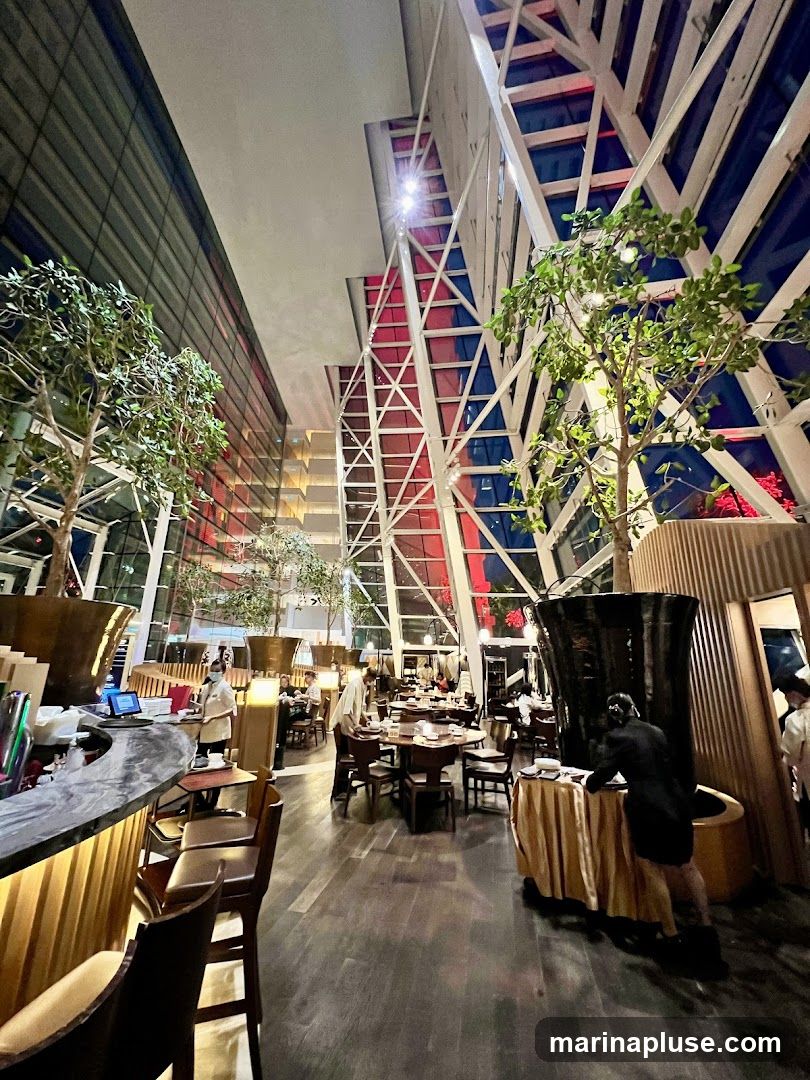 A spacious restaurant interior with a modern, architectural design. The space features a high ceiling with a striking geometric structure, large windows, and ambient lighting. Several trees are placed throughout the dining area, adding a touch of nature. Tables are set for diners, and staff members are visible attending to guests and preparing for service. The overall atmosphere appears elegant and contemporary.