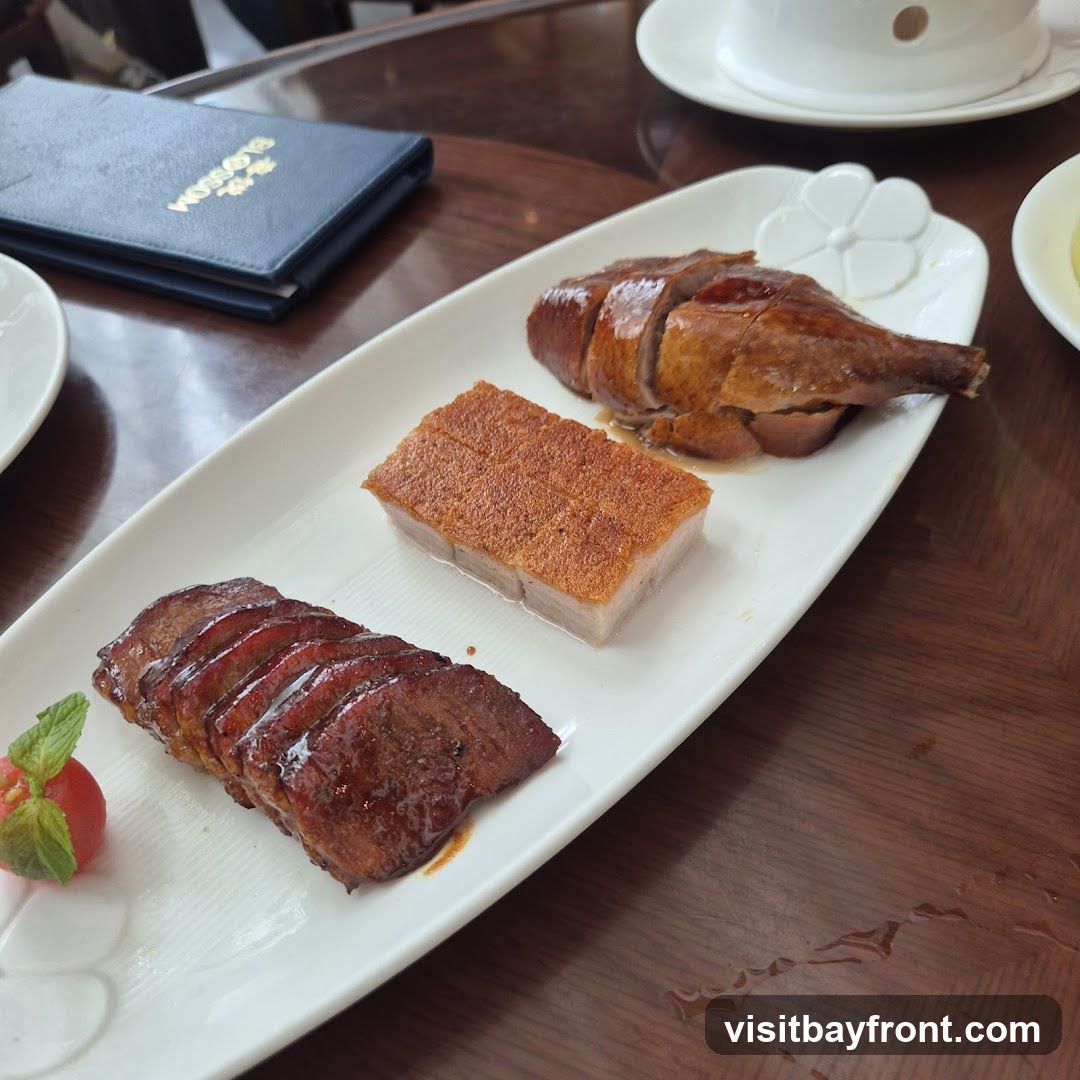 A white platter holds three types of roasted meat: sliced char siu, crispy pork belly, and roasted duck. The char siu is glazed and appears tender. The pork belly has a golden-brown, crackling skin. The roasted duck is sliced and has a glossy finish. A small garnish of mint leaves and a cherry tomato sits next to the char siu. A dark blue menu with gold lettering is visible in the background. The platter is on a wooden table.
