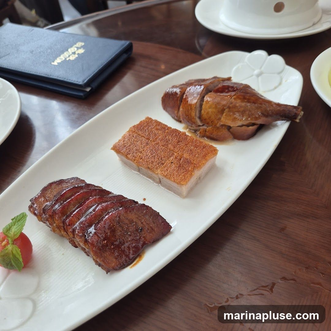 A white platter holds three types of roasted meat: sliced char siu, crispy pork belly, and roasted duck. The char siu is glazed and appears tender. The pork belly has a golden-brown, crackling skin. The roasted duck is sliced and has a glossy finish. A small garnish of mint leaves and a cherry tomato sits next to the char siu. A dark blue menu with gold lettering is visible in the background. The platter is on a wooden table.