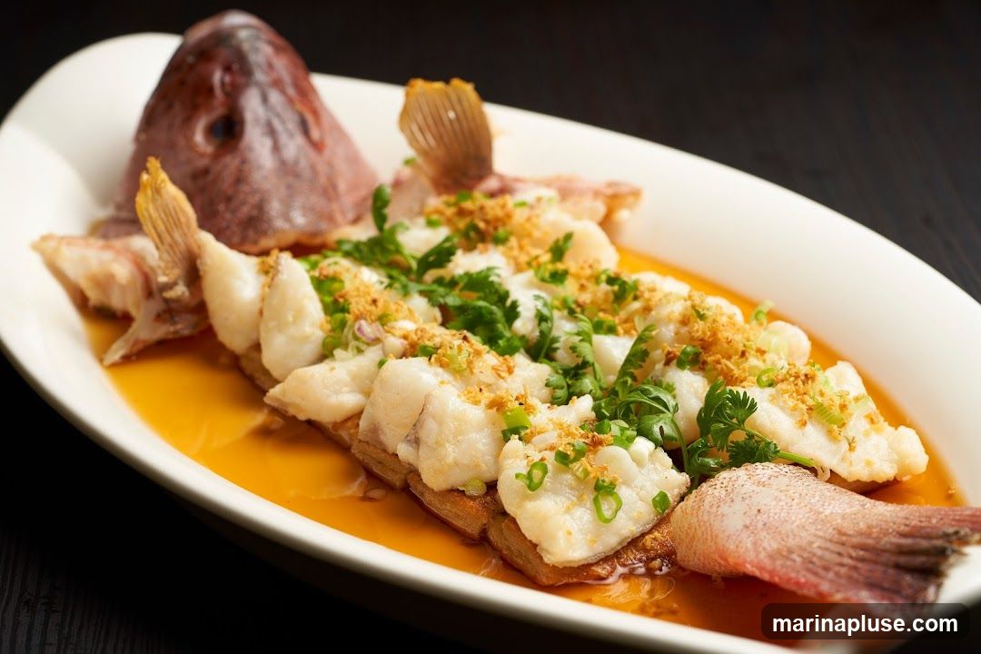 A whole steamed fish, with its head and tail intact, is presented on a white oval platter. The fish is deboned and flaked, arranged over a bed of what appears to be crispy fried wonton wrappers or similar base. It is generously garnished with chopped scallions, fresh cilantro, and crispy fried garlic bits. The fish is bathed in a savory, amber-colored sauce. The dark background contrasts with the white platter and the vibrant colors of the dish.