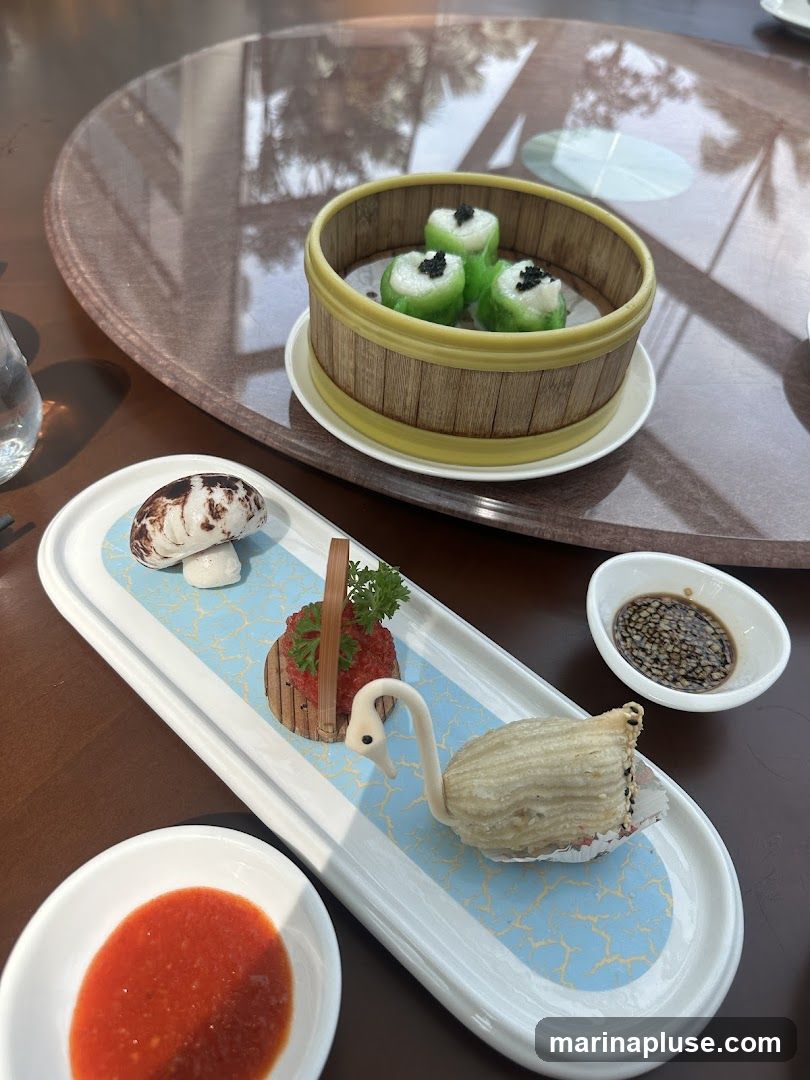 A close-up shot of a dim sum platter featuring various delicate dishes. In a bamboo steamer, there are three green dumplings topped with black caviar. A long white platter displays a mushroom-shaped pastry, a small basket with red roe and parsley, and a fried pastry shaped like a swan. Two dipping sauces, one red and one dark brown, are served alongside. The dishes are arranged on a polished wooden table with reflections of palm trees and sunlight.