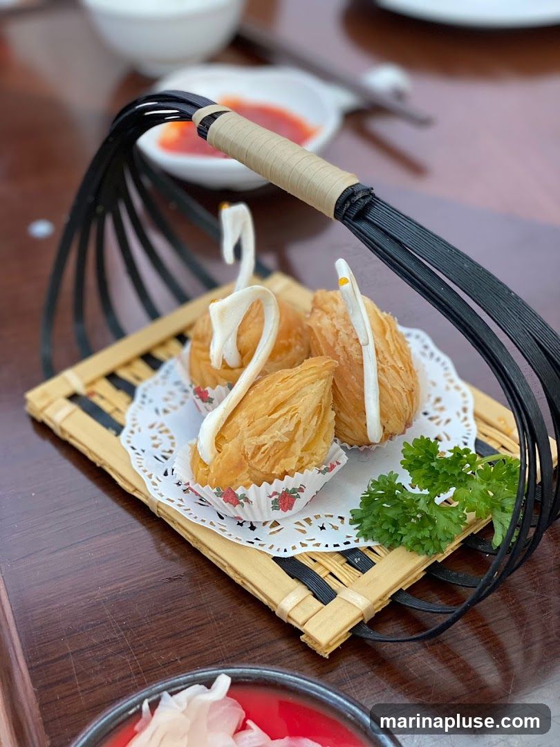 Three golden-brown pastries shaped like swans are presented in a black wire basket lined with a bamboo mat and a doily. Each pastry is in a decorative paper cup and garnished with a white frosting beak and eyes. A sprig of parsley adds a touch of green. In the background, out of focus, are bowls of sauce and what appears to be a white ceramic dish.