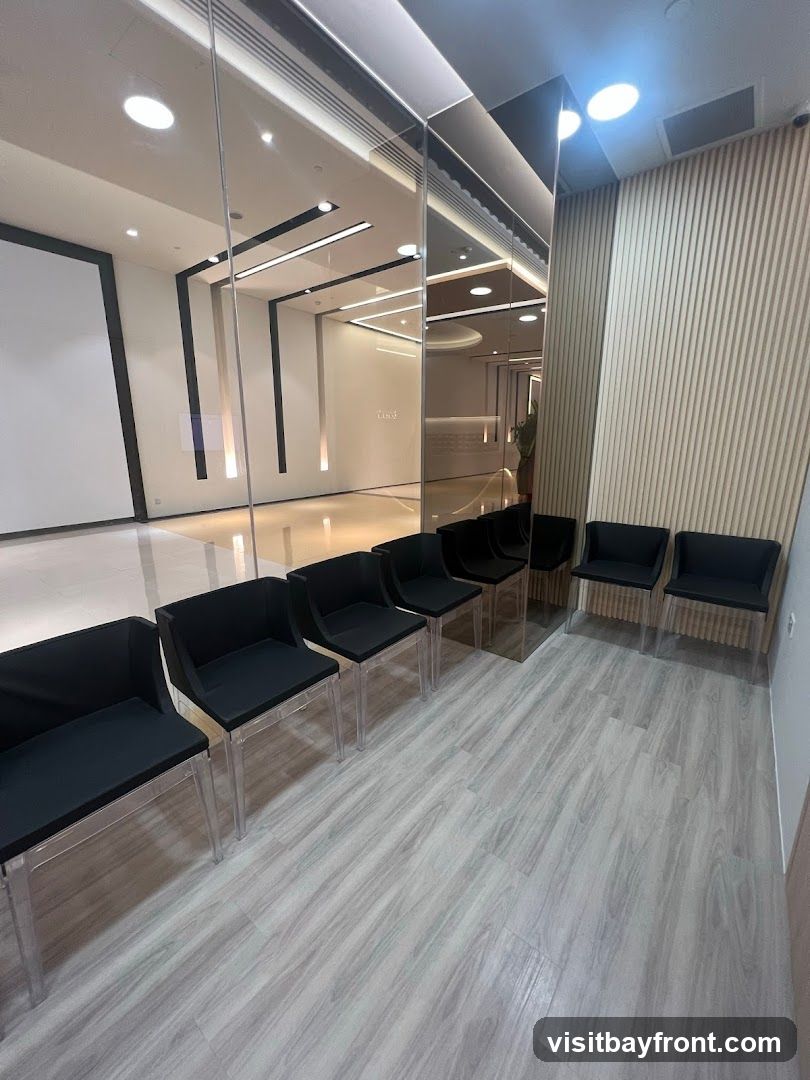 A clean and modern waiting area with a row of black chairs with clear legs, arranged on a light wood-look floor. The space features a mirrored wall reflecting the interior, which includes sleek lighting and a large blank screen or projection surface. The opposite wall is paneled with vertical wooden slats.