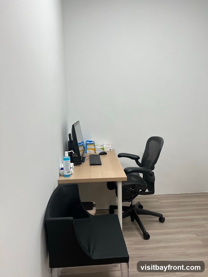 A clean and minimalist medical office setting featuring a wooden desk with a computer monitor, keyboard, and mouse. A bottle of hand sanitizer and some medical supplies are neatly arranged on the desk. An ergonomic black office chair is positioned behind the desk, and a black visitor chair is placed in front. The walls are plain white, and the floor has a light wood laminate finish.