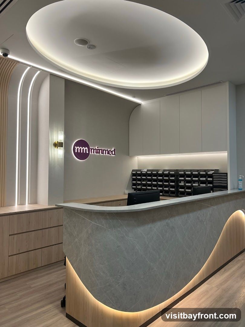 A modern reception area with a curved counter. The counter has a marble-like finish and is illuminated with warm LED lighting. Behind the counter, there are rows of mail slots and a computer monitor. To the left, there is a wall with vertical LED strip lighting and a circular logo that reads 'mm minmed'. Wooden cabinets are visible on the left side of the image.