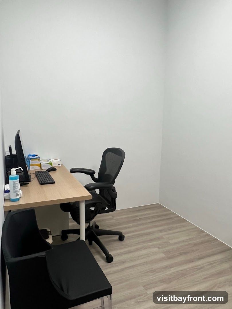 A clean and minimalist office room with a desk, two chairs, and a computer. The room has white walls and light wood-look flooring. On the desk, there is a computer monitor, keyboard, mouse, a bottle of hand sanitizer, and some boxes of medical supplies. One chair is an ergonomic office chair, and the other is a black chair with a clear plastic base.