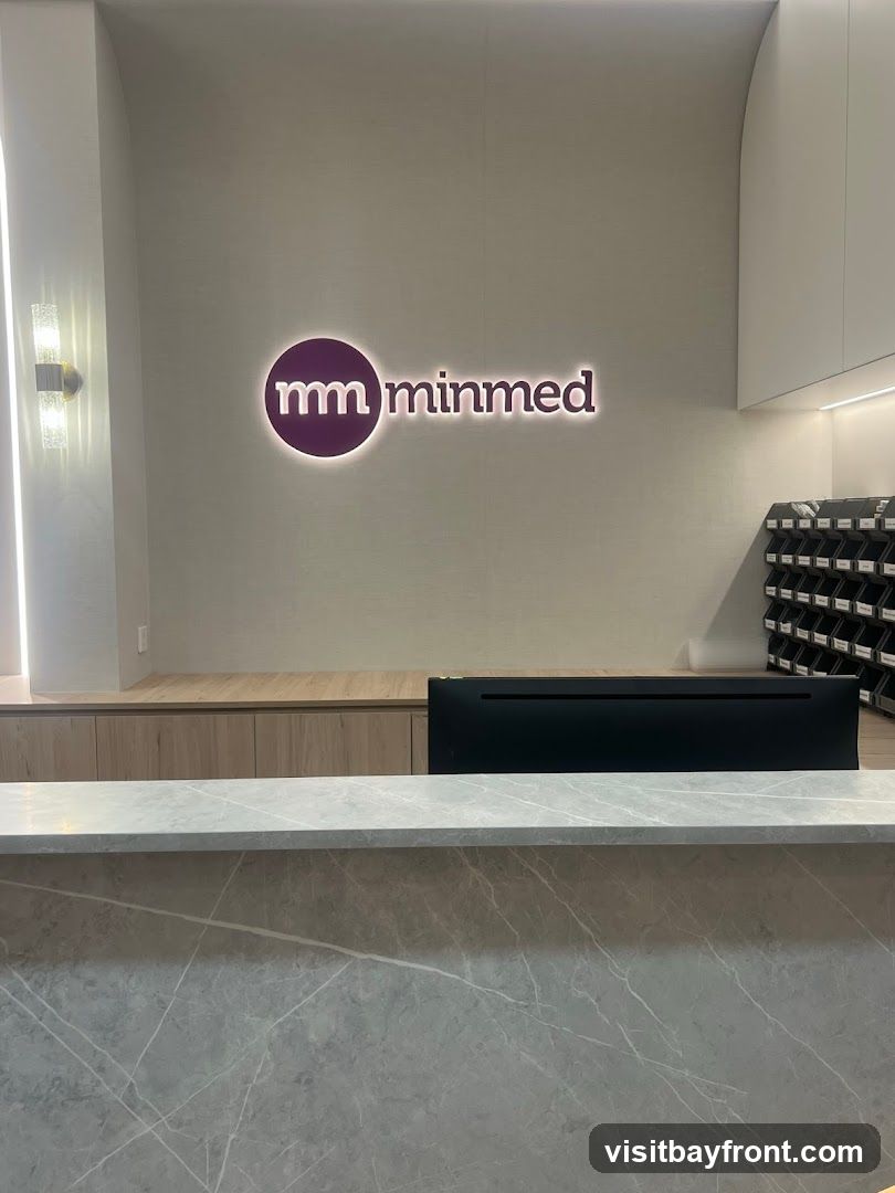 A reception desk with a marble-patterned front is in the foreground. Behind the desk, a wall features a glowing purple logo with the letters "mm" inside a circle, followed by the word "minmed" in illuminated text. To the right, a wall-mounted organizer with many small black compartments is visible. A computer monitor sits on the desk.