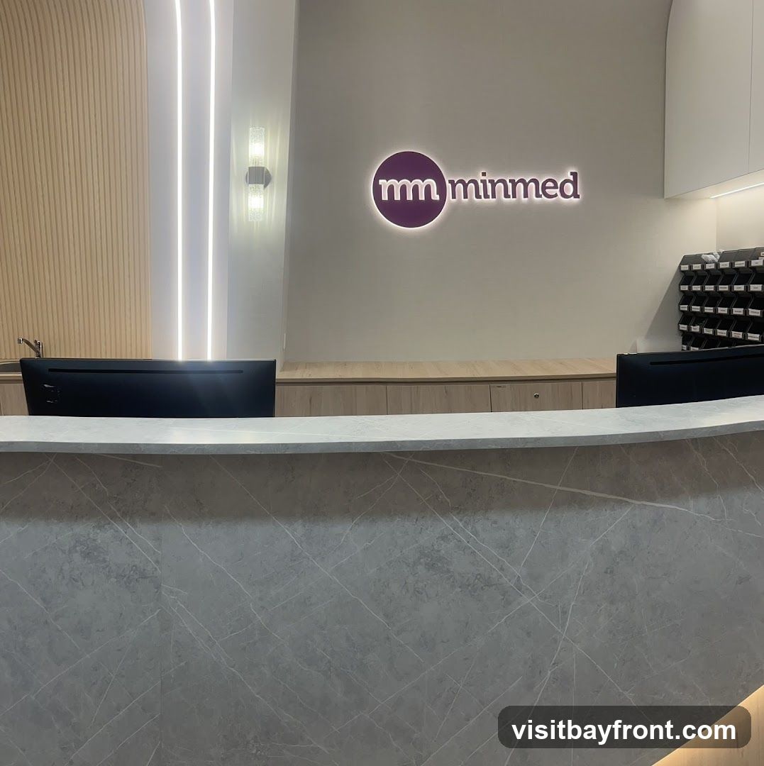 A modern reception area with a curved counter in the foreground. Behind the counter, a wall features a glowing logo that reads "mm minmed". To the left, vertical wooden panels and strip lighting create a contemporary aesthetic. To the right, a wall-mounted organizer with multiple compartments is visible. Two computer monitors are placed on the counter.