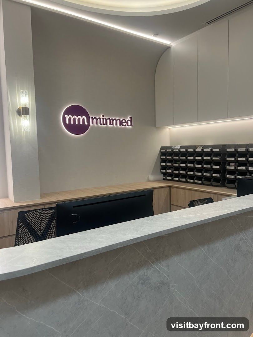 A modern reception area with a prominent logo on the wall. The logo features a purple circle with "mm" inside, followed by the word "minmed" in white lettering. To the right of the logo, there is a wall-mounted organizer with many small compartments. A computer monitor is visible on the reception desk, which is made of a light-colored marble-like material. The overall ambiance is clean and professional.