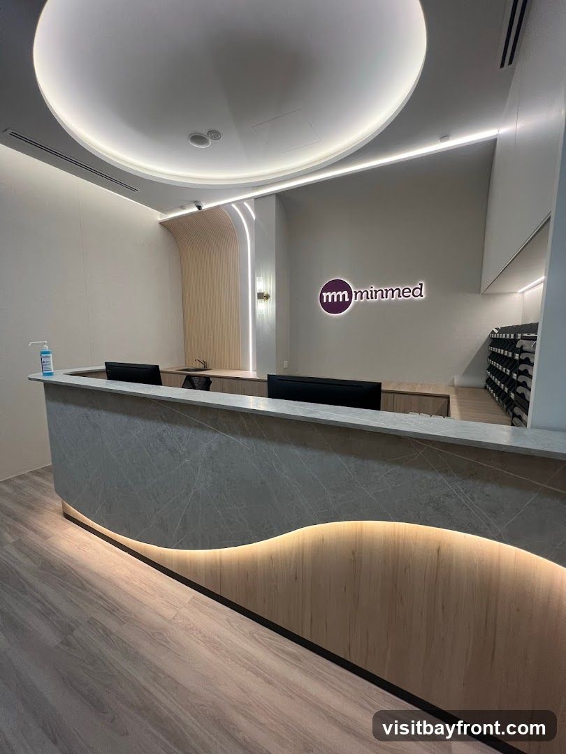 A modern reception area with a curved counter. The counter has a grey marble-like finish on the front and a wood-grain finish on the bottom, illuminated by soft lighting. Behind the counter, a logo with the text "mm minmed" is visible on the wall. The ceiling features a large circular light fixture.