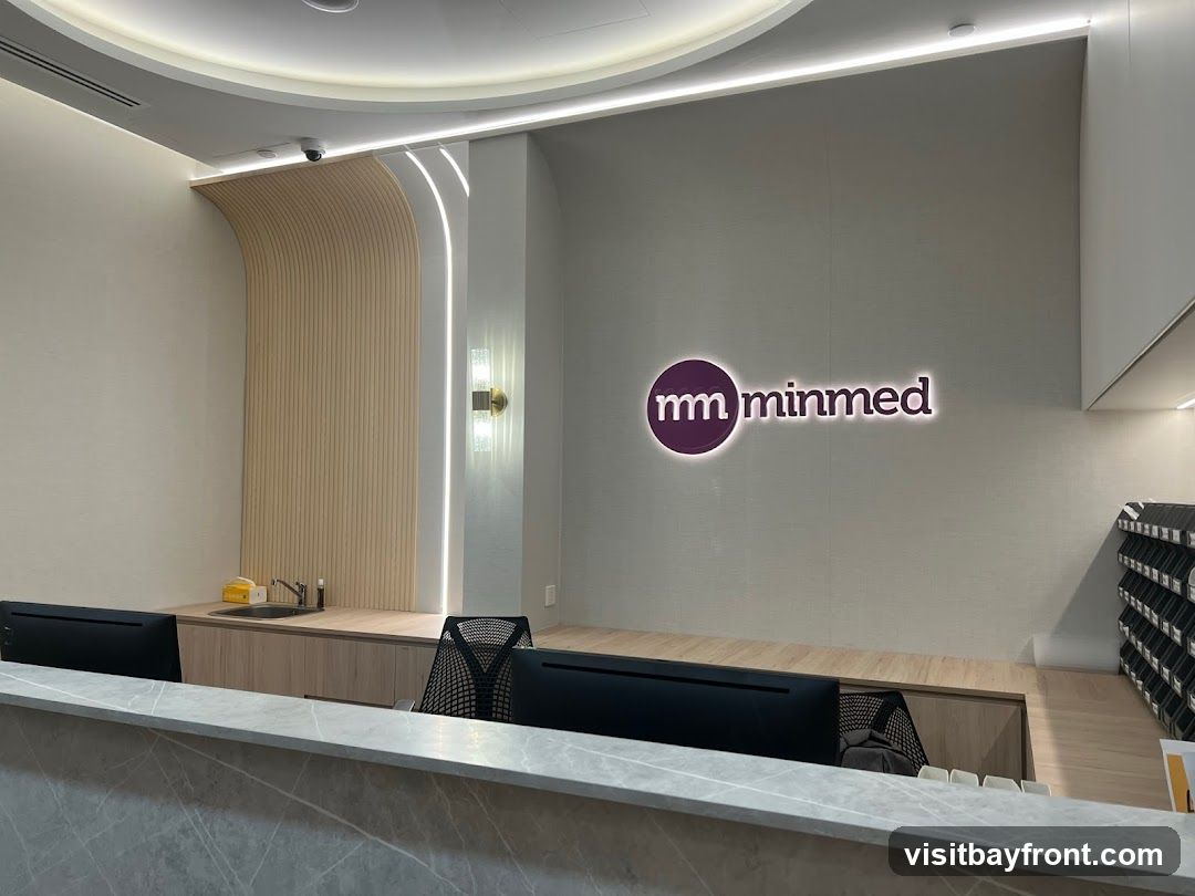 A modern and clean reception area with the Minmed logo prominently displayed on the wall. The reception desk has computers and a marble countertop. Behind the desk, there is a sink area and a wall with vertical wood paneling and accent lighting. To the right, there are shelves with what appear to be medical supplies or patient files.