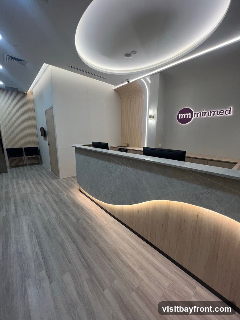 A sleek and modern reception area with a curved counter, wood paneling, and ambient lighting. The 'minmed' logo is visible on the wall. Two computers are on the counter. In the background, there is a waiting area with chairs and a door.