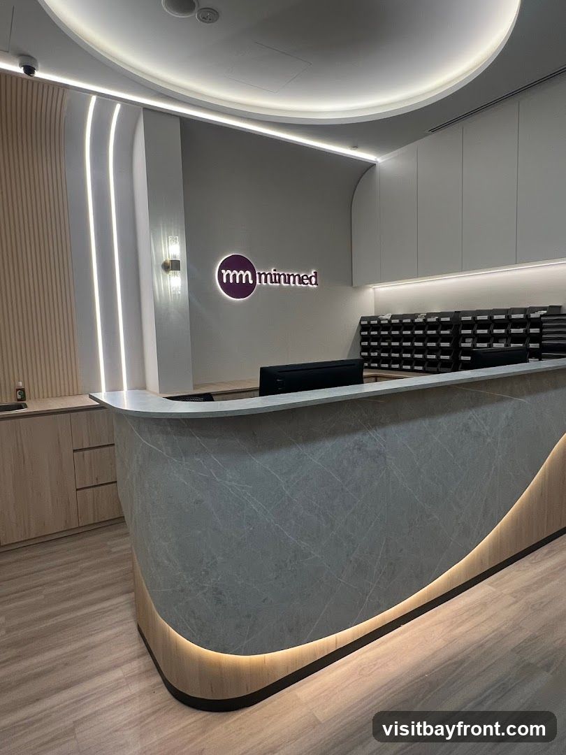 A modern reception area with a curved counter. The counter has a grey marble-like finish with a wooden base illuminated by warm LED lights. Behind the counter, there is a wall with the logo "mm minmed" and a series of black filing trays. The floor is made of light-colored wood-look planks.
