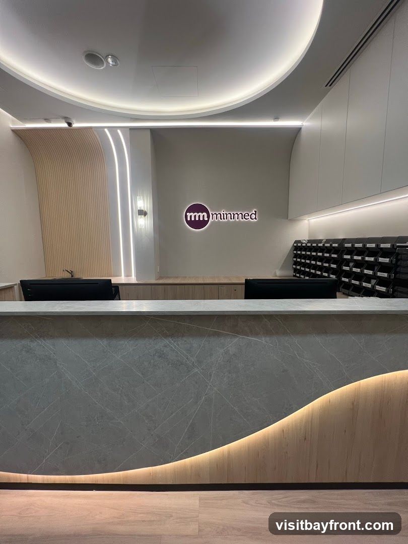 A modern reception area with a marble-patterned counter, wooden accents, and soft lighting. A logo with the text "mm minmed" is visible on the wall. There are two computer monitors on the counter and a mail sorter behind it.