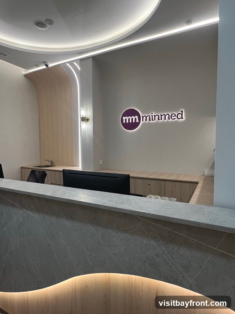 A modern reception area with a counter in the foreground and a wall with a logo and lighting in the background. The logo reads 'mm minmed'. There is a computer monitor on the counter and a sink area behind it.