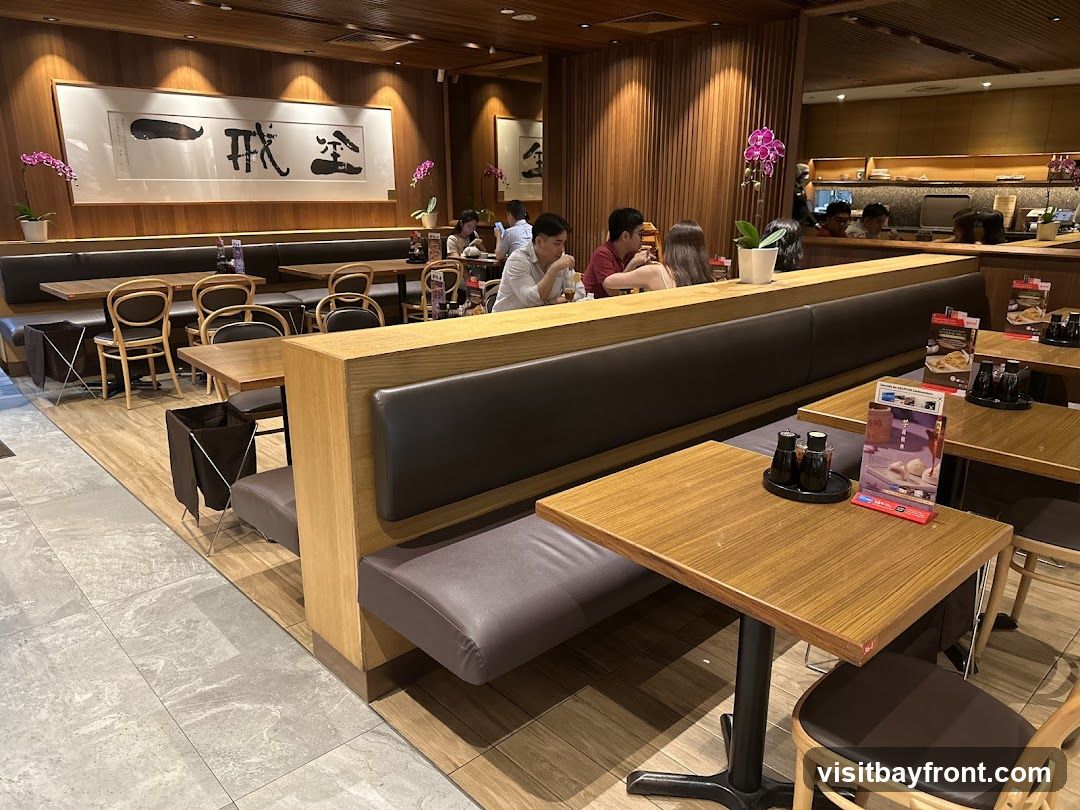 The image shows the interior of a restaurant with wooden booths and tables. Several diners are seated at the booths, some engaged in conversation. Decorative orchids are placed on the tables and near the booths. A large framed artwork with calligraphy hangs on the wall above the booths. Table settings include condiments and small menu displays. The overall ambiance is warm and inviting, with wood paneling and soft lighting.