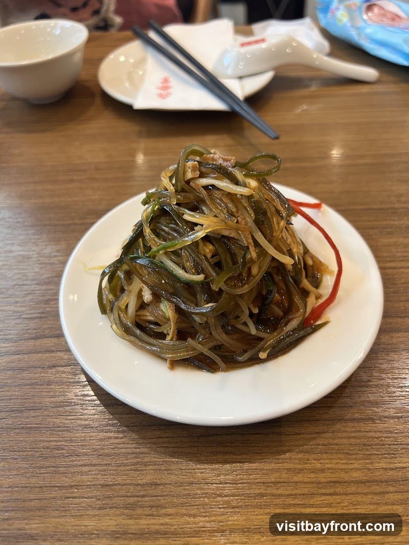 A plate of seaweed salad with bean sprouts and a red garnish sits on a wooden table. Chopsticks and a spoon are visible in the background, along with a white bowl and a napkin with a red logo.