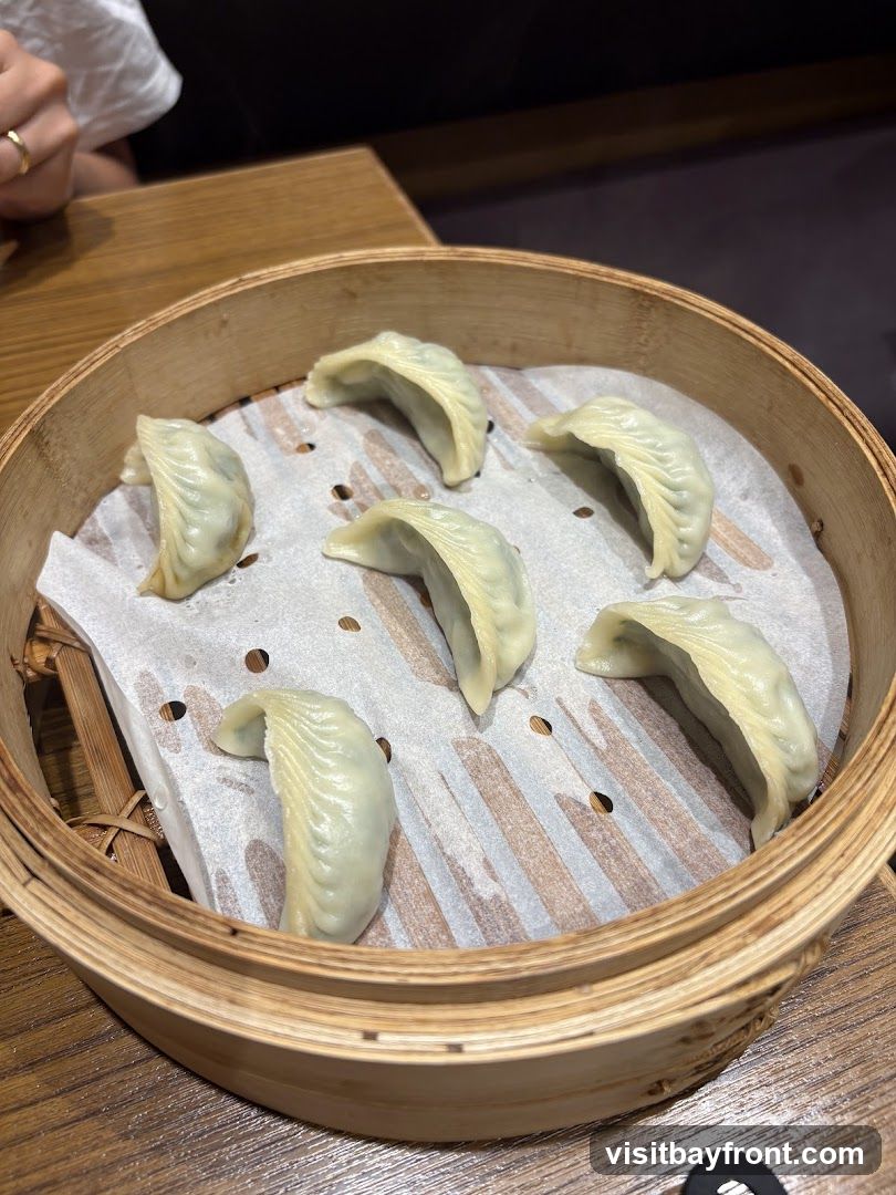 A close-up shot shows six steamed dumplings arranged in a bamboo steamer lined with white parchment paper. The dumplings are pale green and have a pleated appearance. The steamer is placed on a wooden table, and a person's hand with a ring is visible in the upper left corner.