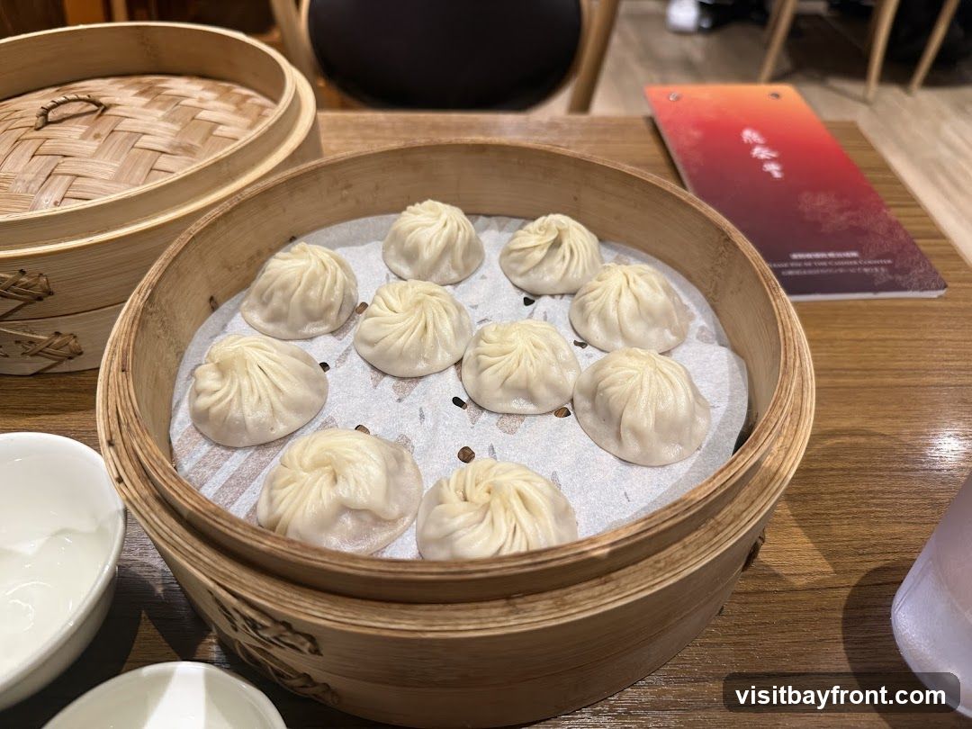 A bamboo steamer basket filled with ten soup dumplings sits on a wooden table. Another bamboo steamer lid is visible in the background. A menu with red and orange gradient and Chinese characters is also on the table, along with a glass of water and small white bowls.