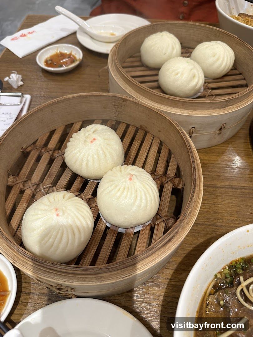 A close-up shot of several bamboo steamers filled with steamed buns, likely soup dumplings, on a wooden table. A small dish of dipping sauce and a spoon are also visible. In the foreground, a bowl of noodle soup with beef and green onions is partially visible.