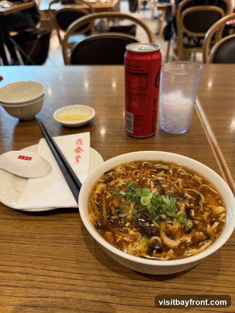 A bowl of hot and sour soup is served on a wooden table, accompanied by a can of Coca-Cola Zero, a glass of ice water, chopsticks, and a small dish of sauce. The soup is garnished with chopped green onions and appears to contain various ingredients such as mushrooms, tofu, and egg. In the background, blurred images of restaurant seating and patrons are visible.