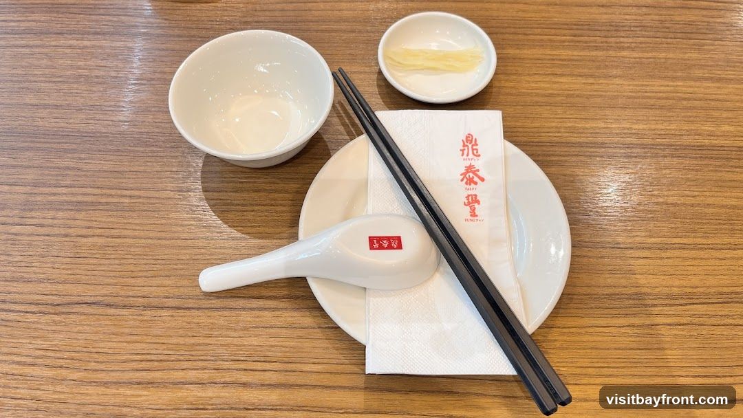 A table setting with a white bowl, a small dish of pickled ginger, a white plate, a white spoon, chopsticks, and a white napkin with red Chinese characters. The setting is on a wooden table.